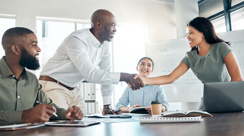 Four smiling colleagues gathered around a meeting table covered with a booklet, phone, coffee cup, day planner, tablet and laptop, with two people seated and two standing as they shake hands